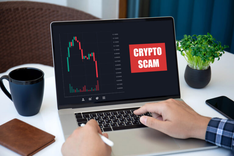 Laptop displaying falling crypto chart and “Crypto Scam” warning beside a digital trading dashboard, illustrating cryptocurrency fraud and Bitcoin investment scams.
