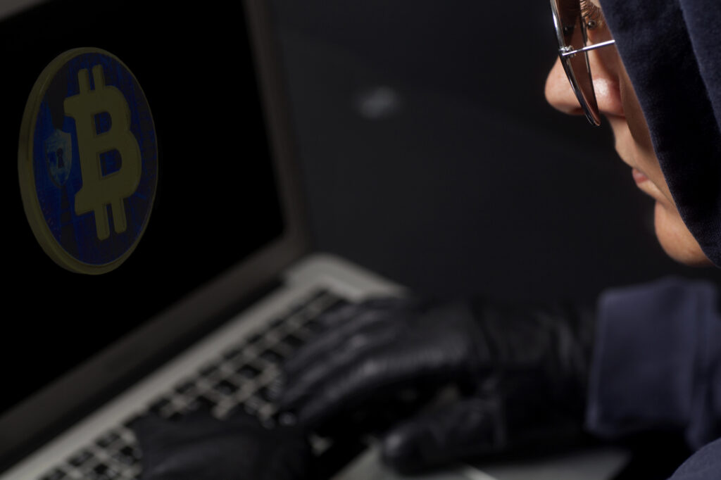 Hooded individual using laptop beside glowing Bitcoin symbol, representing online cryptocurrency fraud, hacking, and digital scam activity.